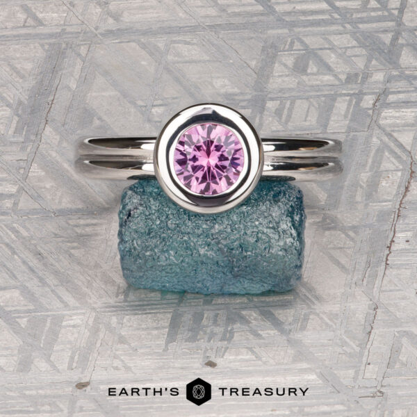 The "Lola" ring in 14k white gold with 0.71-carat Montana sapphire The "Lola" ring in 14k white gold, featuring a 0.71-carat round Montana sapphire, is displayed upright on a rough blue stone with a geometric background and the Earth's Treasury logo at the bottom.
