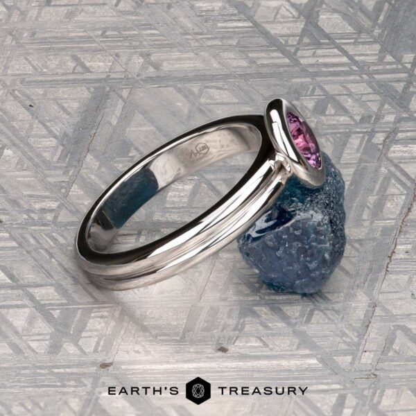 The "Lola" ring in 14k white gold with 0.71-carat Montana sapphire The "Lola" ring in 14k white gold with a 0.71-carat Montana sapphire rests on a rough blue stone atop a patterned metallic surface. "EARTH'S TREASURY" appears at the bottom.