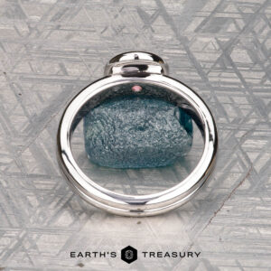 The "Lola" ring, crafted in 14k white gold and set with a 0.71-carat Montana sapphire, is showcased on a geometric-patterned surface with the text "EARTH'S TREASURY" below.
