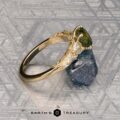 The "Chrysanthemum" ring in 14k yellow gold, featuring a 1.46-carat Australian sapphire and clear accent stones, is displayed on a rough blue crystal atop a patterned metallic surface with the "EARTH'S TREASURY" logo below.