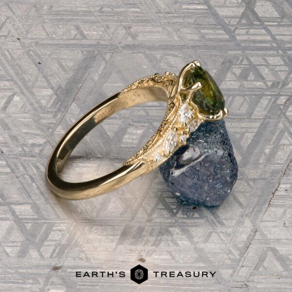 The "Chrysanthemum" ring in 14k yellow gold with 1.46-carat Australian sapphire The "Chrysanthemum" ring in 14k yellow gold, featuring a 1.46-carat Australian sapphire and clear accent stones, is displayed on a rough blue crystal atop a patterned metallic surface with the "EARTH'S TREASURY" logo below.