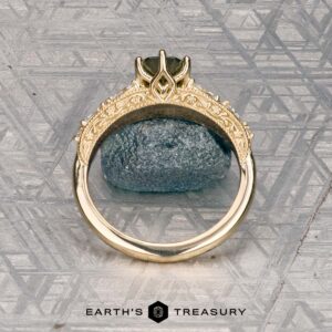 The "Chrysanthemum" ring in 14k yellow gold, featuring a 1.46-carat Australian sapphire, is displayed upright before a textured blue stone on a patterned metallic surface. Logo reads "Earth's Treasury.