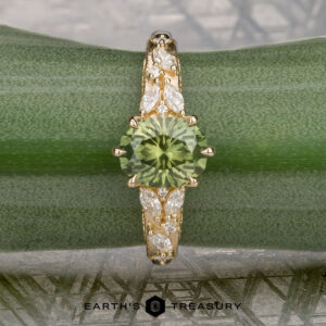 The "Chrysanthemum" ring in 14k yellow gold, featuring a 1.46-carat Australian sapphire and marquise-cut white accent stones, is displayed on a green plant leaf with the "Earth's Treasury" logo below.