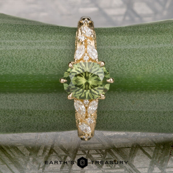 The "Chrysanthemum" ring in 14k yellow gold with 1.46-carat Australian sapphire The "Chrysanthemum" ring in 14k yellow gold, featuring a 1.46-carat Australian sapphire and marquise-cut white accent stones, is displayed on a green plant leaf with the "Earth's Treasury" logo below.