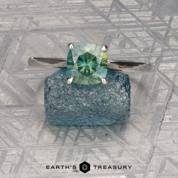 The Cathedral "Fleur" ring in platinum with 1.72-carat Montana sapphire The Cathedral "Fleur" platinum ring with a 1.72-carat Montana sapphire is displayed on a rough blue stone, set against a textured metallic background with geometric patterns. The Earth's Treasury logo appears at the bottom.