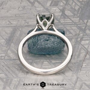 The Cathedral "Fleur" ring in platinum features a 1.72-carat Montana sapphire in an intricate prong setting, displayed upright on a textured blue-gray stone background. Logo text at the bottom reads "Earth's Treasury.
