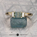 The "Athena" ring in 14K yellow gold, featuring a 1.20-carat Montana sapphire and two baguette diamonds, is displayed on a rough blue stone with a textured background. The Earth’s Treasury logo appears below.