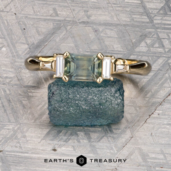 The "Athena" ring in 14K yellow gold with 1.20-carat Montana sapphire The "Athena" ring in 14K yellow gold, featuring a 1.20-carat Montana sapphire and two baguette diamonds, is displayed on a rough blue stone with a textured background. The Earth’s Treasury logo appears below.