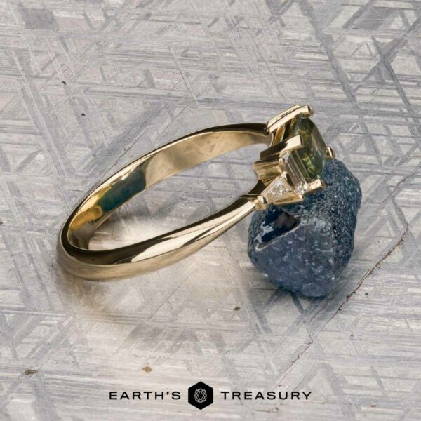 The "Athena" ring in 14K yellow gold with 1.20-carat Montana sapphire The "Athena" ring in 14K yellow gold, featuring a 1.20-carat Montana sapphire, is displayed atop a rough blue-gray mineral on a textured metallic surface.