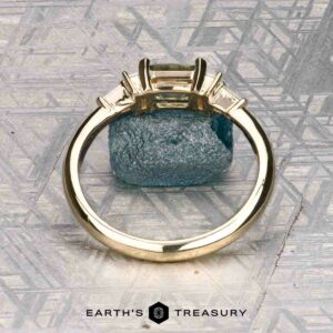 The "Athena" ring in 14K yellow gold with a 1.20-carat Montana sapphire stands upright against a blue mineral and geometric background, with the "EARTH'S TREASURY" logo at the bottom.