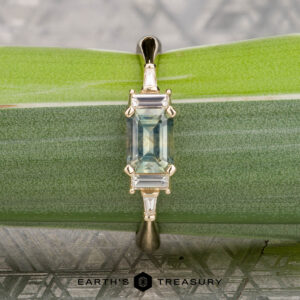 The "Athena" ring in 14K yellow gold features a 1.20-carat rectangular Montana sapphire and baguette side stones, shown on a green leaf with "EARTH'S TREASURY" at the bottom.