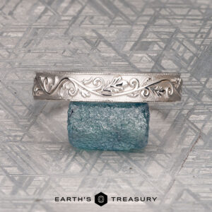 The "Oakley" ring in 14K white gold displayed on a textured blue glass block, set against a geometric patterned background. Earth's Treasury logo at the bottom.