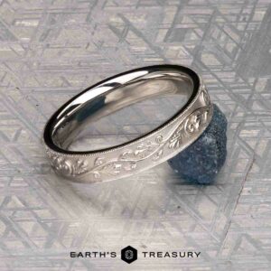 The "Oakley" ring in 14K white gold, engraved with a floral pattern, rests atop a rough blue gemstone on a textured surface, with "Earth's Treasury" displayed below.