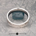 The "Oakley" ring in 14K white gold, featuring engraved designs around a rough blue gemstone, is shown on a metallic textured surface with "EARTH'S TREASURY" at the bottom.