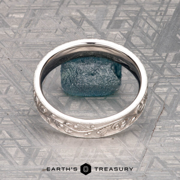 The "Oakley" ring in 14K white gold The "Oakley" ring in 14K white gold, featuring engraved designs around a rough blue gemstone, is shown on a metallic textured surface with "EARTH'S TREASURY" at the bottom.