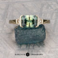 The "Dinara" ring in 14K yellow gold, featuring a 1.72-carat Australian sapphire and two side diamonds, stands upright on a rough blue stone atop a textured gray surface.