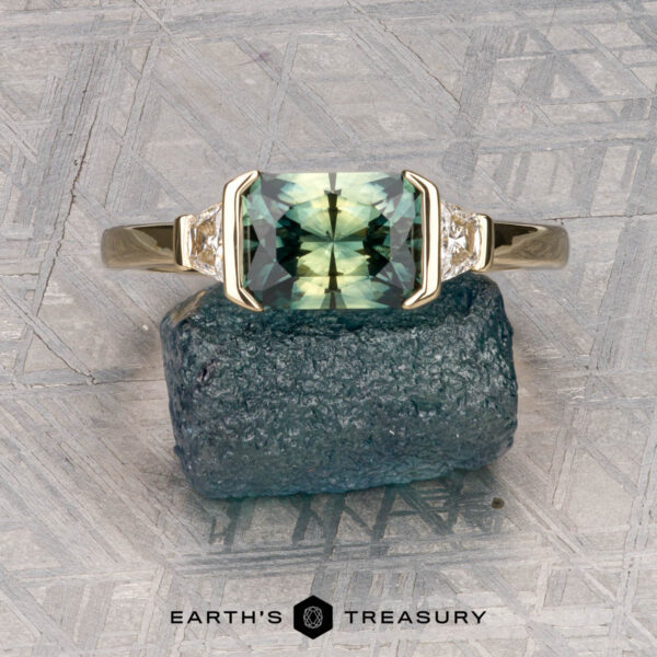 The "Dinara" ring in 14K yellow gold with 1.72-carat Australian sapphire The "Dinara" ring in 14K yellow gold, featuring a 1.72-carat Australian sapphire and two side diamonds, stands upright on a rough blue stone atop a textured gray surface.