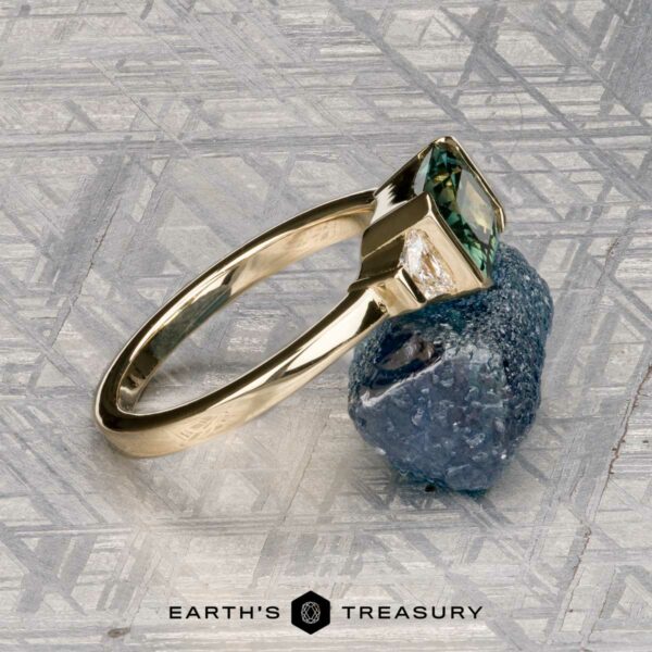 The "Dinara" ring in 14K yellow gold with 1.72-carat Australian sapphire The "Dinara" ring in 14K yellow gold features a 1.72-carat Australian sapphire and a rough blue stone, displayed on a patterned metallic surface with the Earth's Treasury logo below.