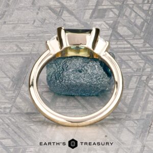The "Dinara" ring in 14K yellow gold, featuring a 1.72-carat Australian sapphire, stands upright on a textured stone background with the Earth’s Treasury logo at the bottom.