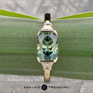 The "Dinara" ring in 14K yellow gold features a 1.72-carat Australian sapphire and two clear side stones, displayed on a green leaf background with "Earth's Treasury" text at the bottom.