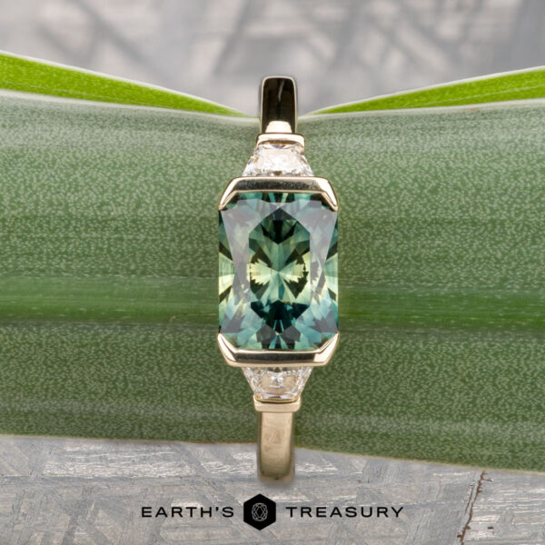 The "Dinara" ring in 14K yellow gold with 1.72-carat Australian sapphire The "Dinara" ring in 14K yellow gold features a 1.72-carat Australian sapphire and two clear side stones, displayed on a green leaf background with "Earth's Treasury" text at the bottom.