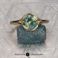The "Brynn" ring in 14K yellow gold with a 1.96-carat sapphire is displayed atop a rough blue stone, set against a geometric patterned background and the "Earth's Treasury" logo below.