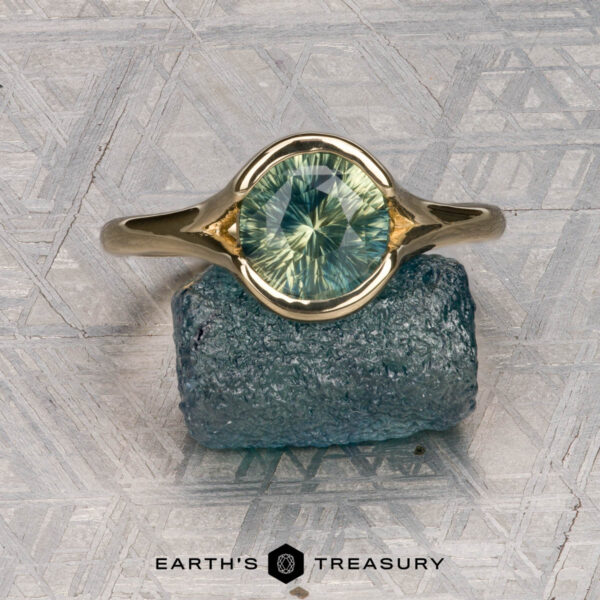 The "Brynn" ring in 14K yellow gold with 1.96-carat sapphire The "Brynn" ring in 14K yellow gold with a 1.96-carat sapphire is displayed atop a rough blue stone, set against a geometric patterned background and the "Earth's Treasury" logo below.