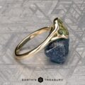 The "Brynn" ring in 14K yellow gold with a 1.96-carat sapphire rests on a rough blue stone atop a patterned metallic surface. The "Earth’s Treasury" logo and name appear at the bottom.