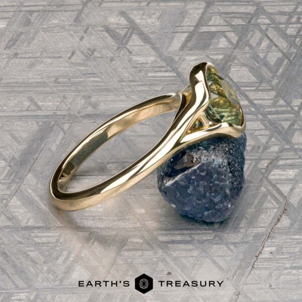 The "Brynn" ring in 14K yellow gold with 1.96-carat sapphire The "Brynn" ring in 14K yellow gold with a 1.96-carat sapphire rests on a rough blue stone atop a patterned metallic surface. The "Earth’s Treasury" logo and name appear at the bottom.