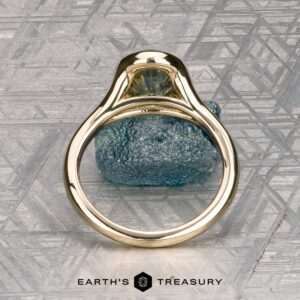 The "Brynn" ring in 14K yellow gold with a 1.96-carat sapphire stands upright on a textured metallic surface, with the "EARTH'S TREASURY" logo displayed at the bottom.
