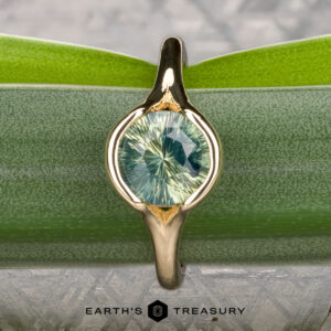 The "Brynn" ring in 14K yellow gold, featuring a 1.96-carat round light green sapphire, is displayed upright on a green leaf with the text "EARTH'S TREASURY" at the bottom.