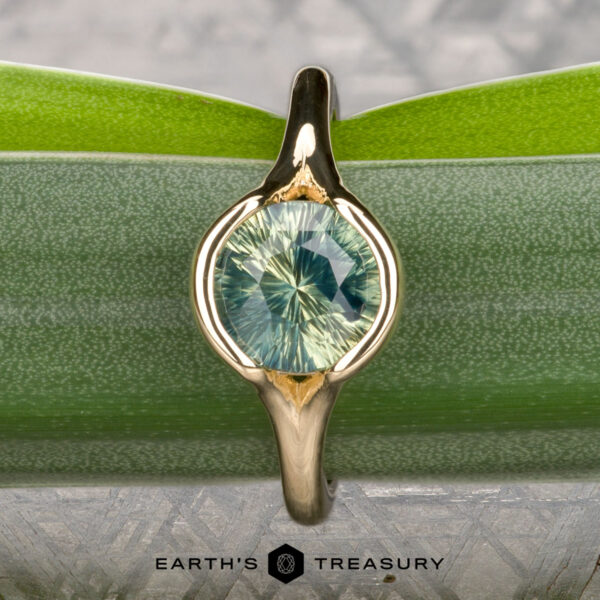 The "Brynn" ring in 14K yellow gold with 1.96-carat sapphire The "Brynn" ring in 14K yellow gold, featuring a 1.96-carat round light green sapphire, is displayed upright on a green leaf with the text "EARTH'S TREASURY" at the bottom.