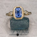 The "Sadie" ring in 14K yellow gold with a 1.56-carat Ceylon sapphire—a rectangular blue center stone flanked by two round white stones—displayed on a rough blue mineral, with the "Earth's Treasury" logo beneath.