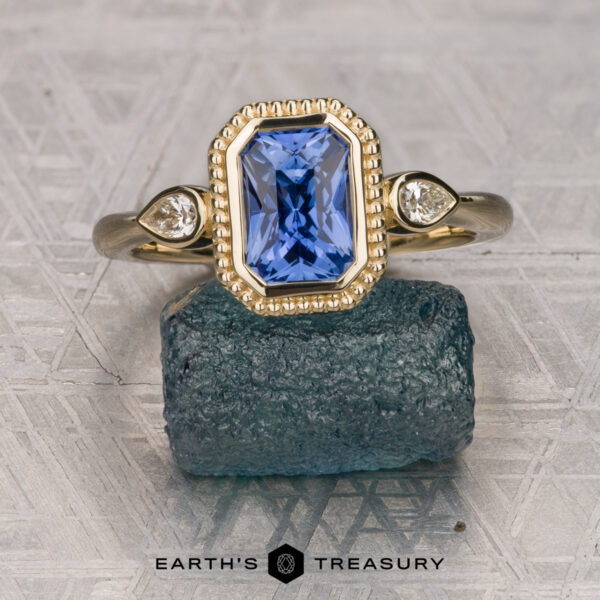 The "Sadie" ring in 14K yellow gold with 1.56-carat Ceylon sapphire The "Sadie" ring in 14K yellow gold with a 1.56-carat Ceylon sapphire—a rectangular blue center stone flanked by two round white stones—displayed on a rough blue mineral, with the "Earth's Treasury" logo beneath.