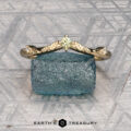 The "Aster" band in 14K yellow gold, featuring a yellow gemstone and leaf details, stands upright on a rough blue stone with the "Earth's Treasury" logo beneath.