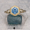 The "Sadie" ring features a 1.49-carat Montana sapphire in a hexagon cut, accented by two small round diamonds and set in 14K yellow gold, displayed on a rough blue stone.