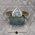 The "Lola" ring in 14k yellow gold with a 1.22-carat Montana sapphire is displayed on a rough blue stone, with the "Earth's Treasury" logo and text below.
