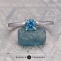 The "Elena" ring in 14K white gold with a 0.93-carat Montana sapphire rests on a raw blue stone, with the "EARTH'S TREASURY" logo visible at the bottom.