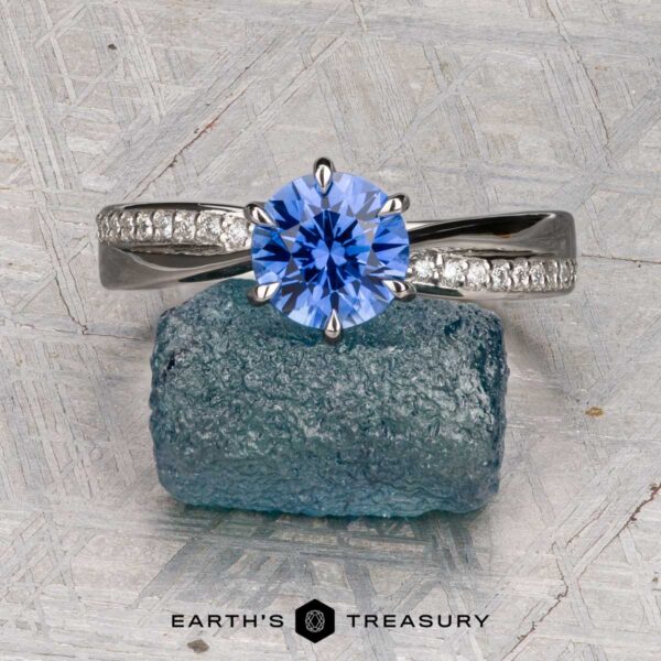 Harari, Moise Harari, Moise: A diamond-accented solitaire silver ring with a round blue gemstone center and delicate diamonds on the band, displayed atop a rough blue stone with "EARTH'S TREASURY" below.