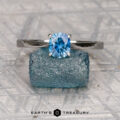 The Cathedral "Fleur" ring in platinum with a 0.95-carat Montana sapphire rests on a rough blue crystal, set against a patterned backdrop with "EARTH'S TREASURY" beneath.