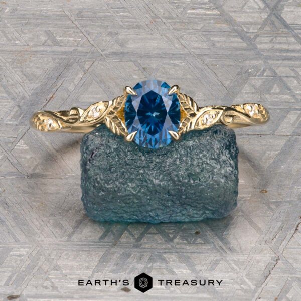 The "Amborella" ring in 14K yellow gold with 0.96-carat Montana sapphire The "Amborella" ring in 14K yellow gold with a 0.96-carat Montana sapphire features delicate leaf engravings and rests on a rough blue-green stone atop a textured surface, with the "Earth's Treasury" logo below.