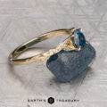 The "Amborella" ring in 14K yellow gold with a 0.96-carat Montana sapphire features delicate leaf engravings and rests on a rough blue-green stone atop a textured surface, with the "Earth's Treasury" logo below.