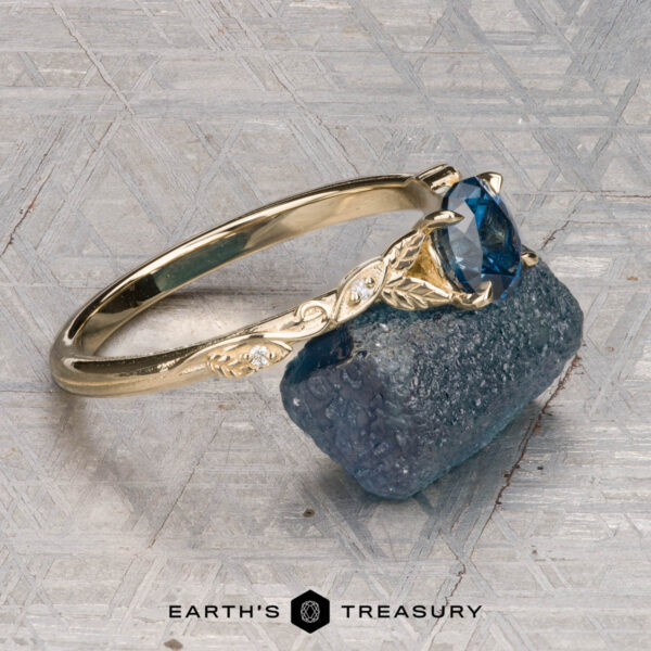 The "Amborella" ring in 14K yellow gold with 0.96-carat Montana sapphire The "Amborella" ring in 14K yellow gold with a 0.96-carat Montana sapphire features delicate leaf engravings and rests on a rough blue-green stone atop a textured surface, with the "Earth's Treasury" logo below.