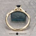 The "Amborella" ring in 14K yellow gold with a 0.96-carat Montana sapphire features delicate leaf engravings and rests on a rough blue-green stone atop a textured surface, with the "Earth's Treasury" logo below.