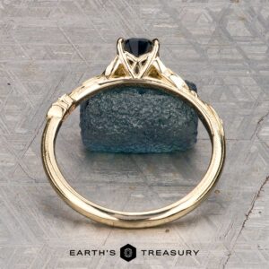 The "Amborella" ring in 14K yellow gold with a 0.96-carat Montana sapphire features delicate leaf engravings and rests on a rough blue-green stone atop a textured surface, with the "Earth's Treasury" logo below.