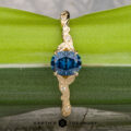 The "Amborella" ring in 14K yellow gold with a 0.96-carat Montana sapphire features delicate leaf engravings and rests on a rough blue-green stone atop a textured surface, with the "Earth's Treasury" logo below.