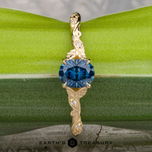 The "Amborella" ring in 14K yellow gold with a 0.96-carat Montana sapphire features delicate leaf engravings and rests on a rough blue-green stone atop a textured surface, with the "Earth's Treasury" logo below.