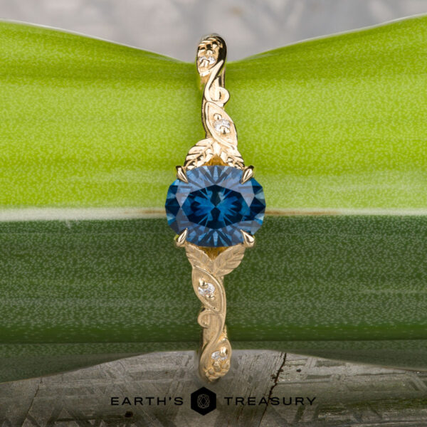 The "Amborella" ring in 14K yellow gold with 0.96-carat Montana sapphire The "Amborella" ring in 14K yellow gold with a 0.96-carat Montana sapphire features delicate leaf engravings and rests on a rough blue-green stone atop a textured surface, with the "Earth's Treasury" logo below.