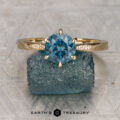 The ring—a gold band with a round blue gemstone and small diamonds—is shown upright on a rough blue stone, with the Earth's Treasury logo below.