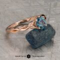 The "Kirryn" ring in 14K rose gold, featuring a 1.17-carat Montana sapphire and leaf motifs, is displayed upright on a rough blue stone background. Earth’s Treasury logo appears at the bottom.